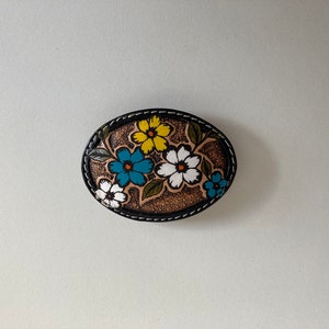 May include: A brown leather belt buckle with a floral design. The flowers are painted in shades of white, yellow, blue, and green.