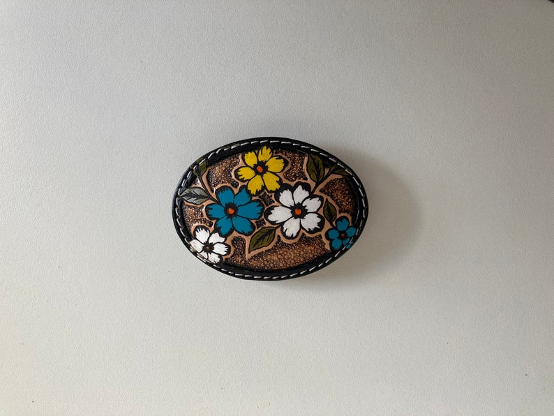 May include: A brown leather belt buckle with a floral design. The flowers are painted in shades of white, yellow, blue, and green.