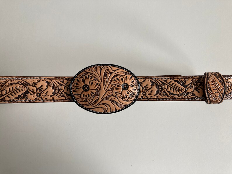 May include: A tan leather belt with a floral design and an oval buckle. The buckle is also decorated with a floral design.