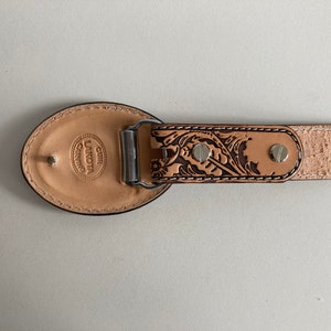 May include: A brown leather belt with a floral design and a silver buckle. The buckle is engraved with the text "CUIR LAKOTA CRAFTING".