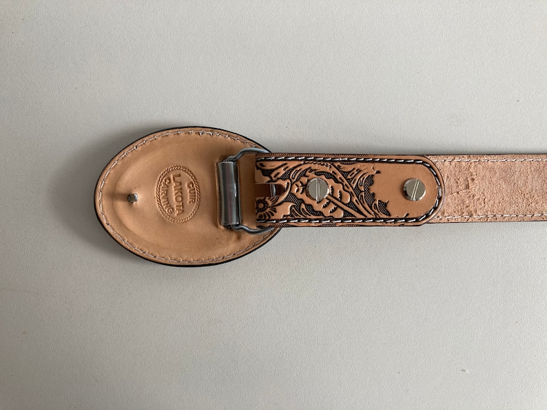 May include: A brown leather belt with a floral design and a silver buckle. The buckle is engraved with the text "CUIR LAKOTA CRAFTING".