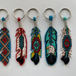 Peut inclure: Cinq porte-clés en cuir en forme de plume colorés avec des anneaux de clés argentés. Chaque porte-clés présente un design différent, y compris des motifs géométriques et une tête d'aigle.