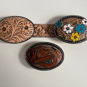 May include: Three leather belt buckles with intricate designs. The first buckle features a floral pattern with two large flowers in the center. The second buckle has a floral pattern with colorful flowers. The third buckle has a dragon design.