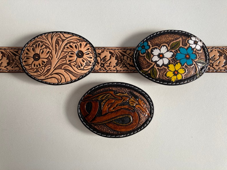 May include: Three leather belt buckles with intricate designs. The first buckle features a floral pattern with two large flowers in the center. The second buckle has a floral pattern with colorful flowers. The third buckle has a dragon design.
