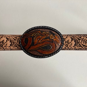 May include: A brown leather belt with a tooled design of flowers and leaves. The belt buckle is oval shaped and features a tooled design of a dragon.