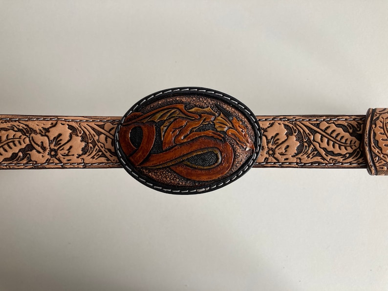 May include: A brown leather belt with a tooled design of flowers and leaves. The belt buckle is oval shaped and features a tooled design of a dragon.