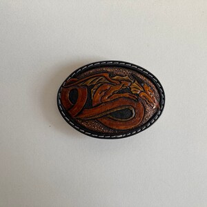 May include: A brown leather belt buckle with a detailed carving of a dragon. The buckle is oval shaped and has a black stitched border.