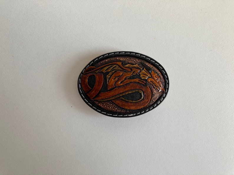 May include: A brown leather belt buckle with a detailed carving of a dragon. The buckle is oval shaped and has a black stitched border.