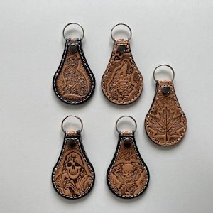 Op de afbeelding: Vijf leren sleutelhangers met verschillende ontwerpen. Elke sleutelhanger heeft een bruine leren hanger met een zwarte gestikte rand en een zilveren sleutelring. De ontwerpen omvatten een wolf, een esdoornblad, een schedel, een kat en een motorfietsmotor.