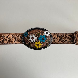 May include: A brown leather belt with a floral design on the buckle. The buckle is oval shaped and has a black border. The flowers are painted in white, yellow, blue, and green.