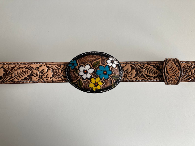 May include: A brown leather belt with a floral design on the buckle. The buckle is oval shaped and has a black border. The flowers are painted in white, yellow, blue, and green.