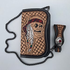 Op de afbeelding: Een bruine leren portemonnee met een zwarte kettingriem en een schedelontwerp. De schedel draagt ​​een rode bandana en heeft een verenhoofdtooi. De portemonnee heeft een geweven patroon en een zilveren sluiting met een rode steen.