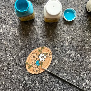 May include: A close-up of a leather craft project with a floral design. The project is being painted with blue acrylic paint using a small paintbrush. There are three bottles of acrylic paint in the background, one white, one blue, and one green.