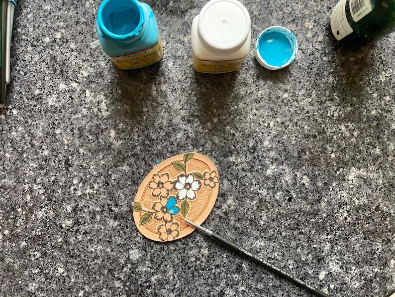 May include: A close-up of a leather craft project with a floral design. The project is being painted with blue acrylic paint using a small paintbrush. There are three bottles of acrylic paint in the background, one white, one blue, and one green.