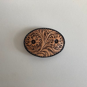 May include: A tan leather belt buckle with a floral design. The buckle is oval shaped and has a black stitched border.
