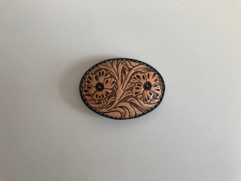 May include: A tan leather belt buckle with a floral design. The buckle is oval shaped and has a black stitched border.