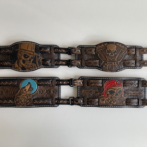 Op de afbeelding: Vier leren armbanden met verschillende ontwerpen. De ontwerpen omvatten een schedel met een hoge hoed, een schedel met een bandana, een wolf die huilt naar de maan en een schedel met een motorfietsmotor.