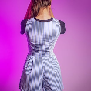 May include: A woman wearing a blue and white striped jumpsuit with a black and white striped trim on the sleeves. The jumpsuit has a wide leg and a relaxed fit.