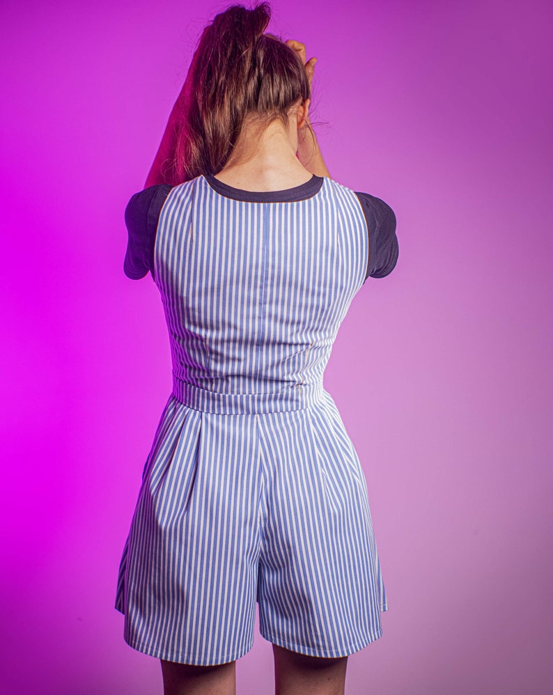 May include: A woman wearing a blue and white striped jumpsuit with a black and white striped trim on the sleeves. The jumpsuit has a wide leg and a relaxed fit.