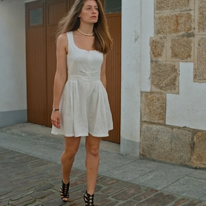 May include: A woman wearing a white linen dress with a pearl necklace and black strappy heels walks on a cobblestone street.