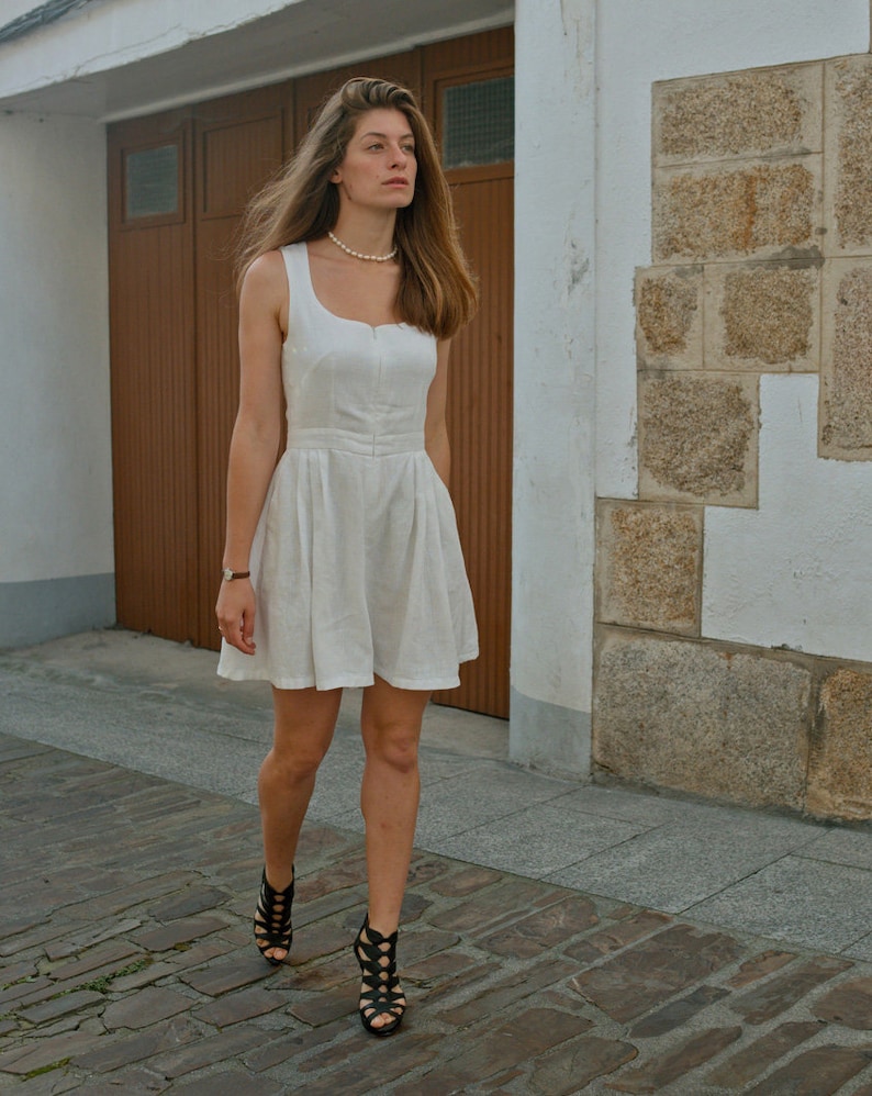 May include: A woman wearing a white linen dress with a pearl necklace and black strappy heels walks on a cobblestone street.