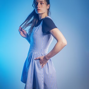 May include: A woman wearing a blue and white striped dress with a black tank top underneath. She is standing in front of a blue background.