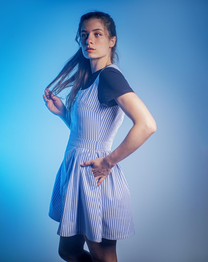 May include: A woman wearing a blue and white striped dress with a black tank top underneath. She is standing in front of a blue background.