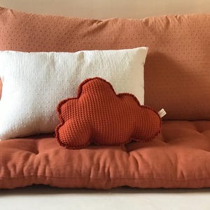 Op de afbeelding: Een wolkvormig decoratief kussen in een warme oranje tint ligt op een bijpassend gecapitonneerd kussen. Een wit rechthoekig kussen met een gestippelde textuur bevindt zich achter het wolkussen. Een groot oranje kussen met een gestippelde textuur staat op de achtergrond.