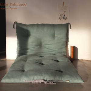 May include: A sage green linen floor cushion with button tufting and a tied closure. The cushion is on a light grey floor.