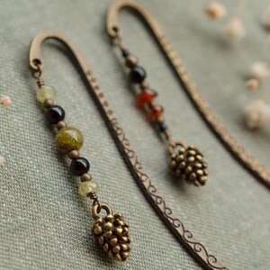Può includere: Due segnalibri in metallo color bronzo con dettagli di perline decorative e un ciondolo a forma di pigna. Un segnalibro presenta perline verdi, nere e gialle. L'altro ha perline rosse, nere e trasparenti. I segnalibri sono su sfondo di tessuto verde.