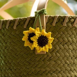 Peut inclure: Gros plan sur un panier vert tissé avec une paire de décorations de tournesols crochetés jaunes et noirs. Le panier a des poignées et des garnitures en cuir clair, suggérant un design naturel et artisanal.