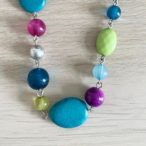 Op de afbeelding: Een kleurrijke ketting met kralen in turquoise, roze, paars, groen, zilver en blauw. De kralen zijn verbonden door zilverkleurige metalen schakels. De ketting heeft een casual, bohemien stijl.