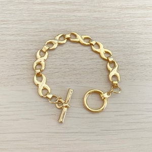 以下が含まれることがあります： ゴールドトーンのブレスレット。無限大の記号のリンクが連なっています。トグルクラスプ留め。明るい木目調の表面に展示されています。普段使いにぴったりのスタイリッシュなアクセサリーです。