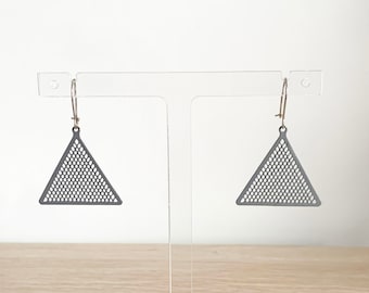 May include: A pair of geometric earrings. Each earring features a dark gray, triangular shape with a perforated design. The earrings hang from gold-colored ear wires. The earrings are displayed on a clear acrylic stand against a white background.