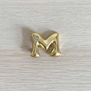 May include: Gold letter M with a clear rhinestone in the center.