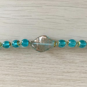 Pode incluir: Um colar de contas de vidro azul turquesa com uma grande conta de vidro em forma de diamante iridescente no centro.
