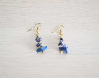 May include: A pair of dangle earrings featuring irregular, deep blue stone chips and small gold-colored beads. The earrings have gold-colored fish hook ear wires. The stones are arranged in a cascading design.