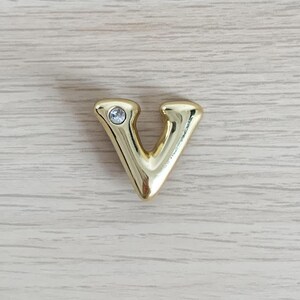 Peut inclure: Une lettre V dorée avec un strass transparent au centre.
