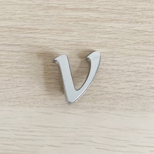 Peut inclure: Une lettre "V" argentée et brillante repose sur une surface en bois clair. La lettre est de style cursif, avec une finition lisse et réfléchissante. Le fond est brun clair, avec un grain de bois visible.