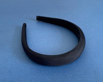 Könnte beinhalten: Ein schwarzes, gepolstertes Stirnband mit glatter, matter Oberfläche. Das Stirnband ist gebogen und hat ein schlichtes, elegantes Design. Das Accessoire befindet sich vor einem blauen Hintergrund.