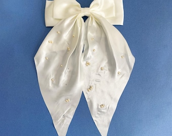 May include: Ivory satin hair bow with a large bow and cascading tails, adorned with scattered faux pearls. The bow is set against a solid blue background, creating a striking contrast. A delicate and elegant hair accessory.
