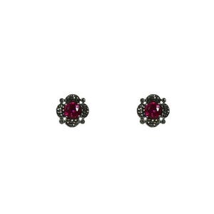 May include: A pair of stud earrings featuring a large, oval, ruby-red gemstone at the center, surrounded by a dark, ornate setting. The setting has a floral design with small, dark accents. The earrings are set against a white background.