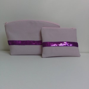 May include: Two lavender-colored cosmetic bags. The larger bag has a curved top and the smaller bag is rectangular. Both bags have a wide band of purple sequins across the front. The bags are zippered.