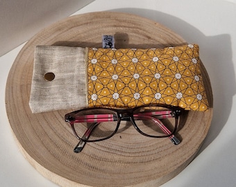 May include: A yellow and white patterned glasses case with a linen flap and a gold button closure. The case is sitting on a wooden surface with a pair of brown glasses on top of it.
