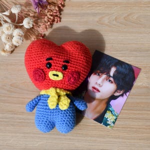 May include: A handmade red heart-shaped crochet doll with a yellow scarf and blue overalls. A photo of a person with dark hair is next to the doll. The doll has black eyes and a yellow mouth.