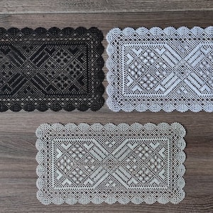 Puede incluir: Tres tapetes rectangulares de encaje en negro, blanco y gris claro, cada uno con un patrón geométrico detallado y un borde festoneado. Los tapetes se exhiben sobre una superficie de madera.