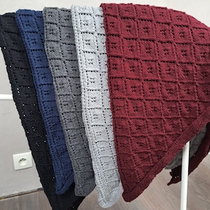 Könnte beinhalten: Eine Präsentation von Strickdecken in verschiedenen Farben, darunter Schwarz, Marineblau, Grau und Burgunderrot. Jede Decke weist ein strukturiertes, quadratisches Muster auf. Die Decken sind über einem weißen Gestell drapiert, um ihre verschiedenen Farbtöne und Texturen zu zeigen.