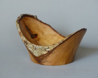 Puede incluir: Un pequeño cuenco de madera con un acabado natural y rústico. El cuenco está hecho de una sola pieza de madera y tiene una forma única y asimétrica. La corteza todavía está unida al borde del cuenco, lo que le da un aspecto natural y orgánico.
