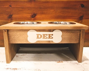 May include: A wooden dog bowl stand with two stainless steel bowls. The stand has a bone-shaped cutout with the name "DEE" engraved on it.
