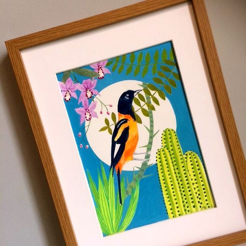 Pu&ograve; includere: Un dipinto incorniciato di un uccello colorato con piume nere, arancioni e gialle appollaiato su un ramo con foglie verdi e fiori rosa. Lo sfondo &egrave; un cielo blu con un cerchio bianco. Ci sono anche piante di cactus verdi nel dipinto.